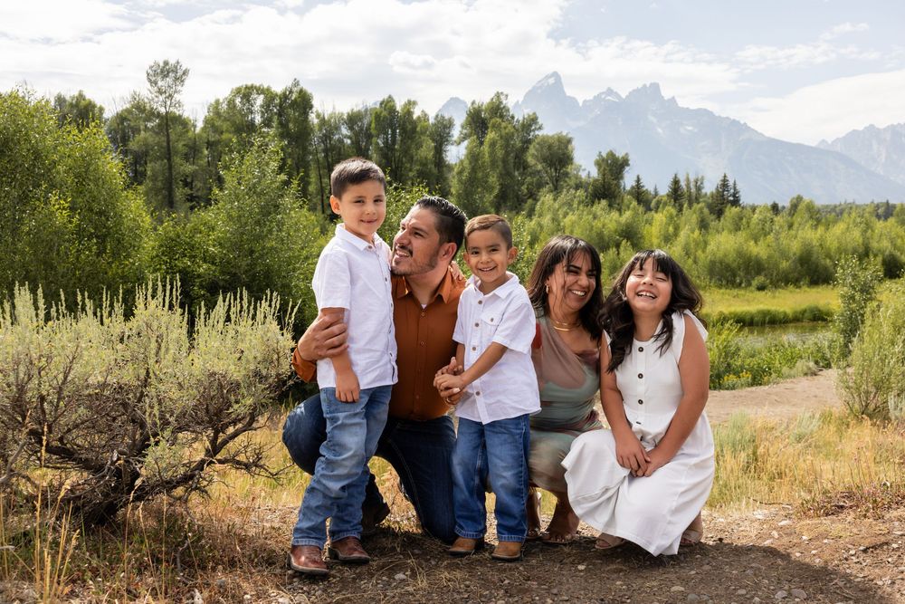 Family of five smiling outdoors with Grand Teton mountains and lush greenery in the background on a sunny day.