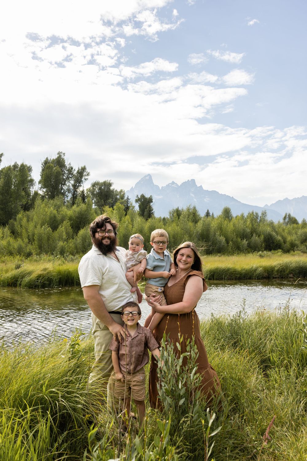 Family of five posing near a mountain stream with Grand Teton peaks and lush greenery in the background.