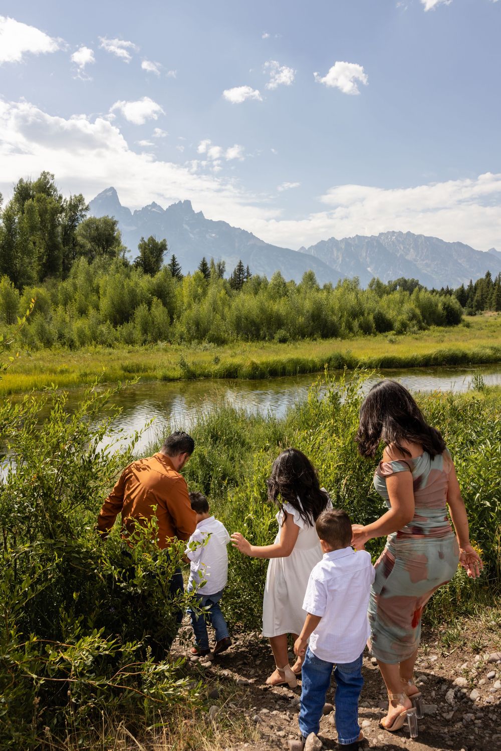 Family of five walking toward a scenic mountain lake surrounded by lush greenery under a bright blue sky.