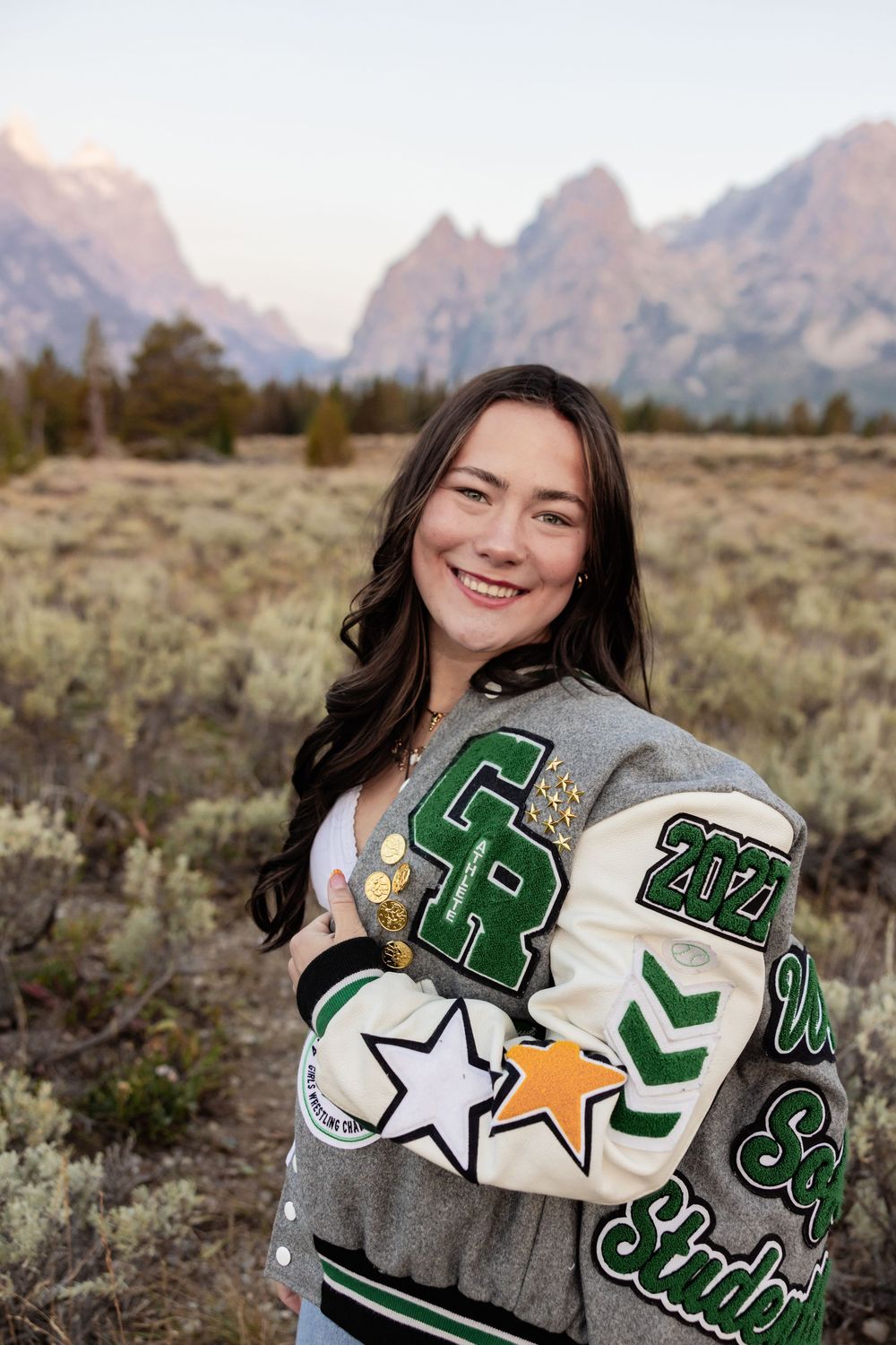 Smiling young woman in a green and white letterman jacket posing in front of the Grand Teton mountains.