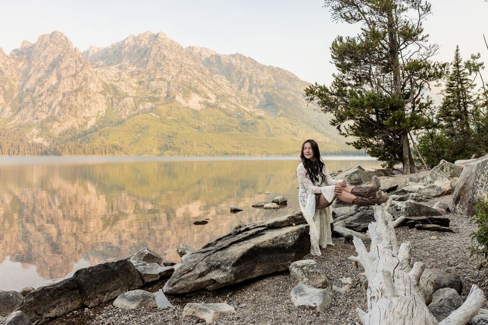 Woman in white dress sitting on rocks by a reflective mountain lake with pine trees at Grand Teton National Park.