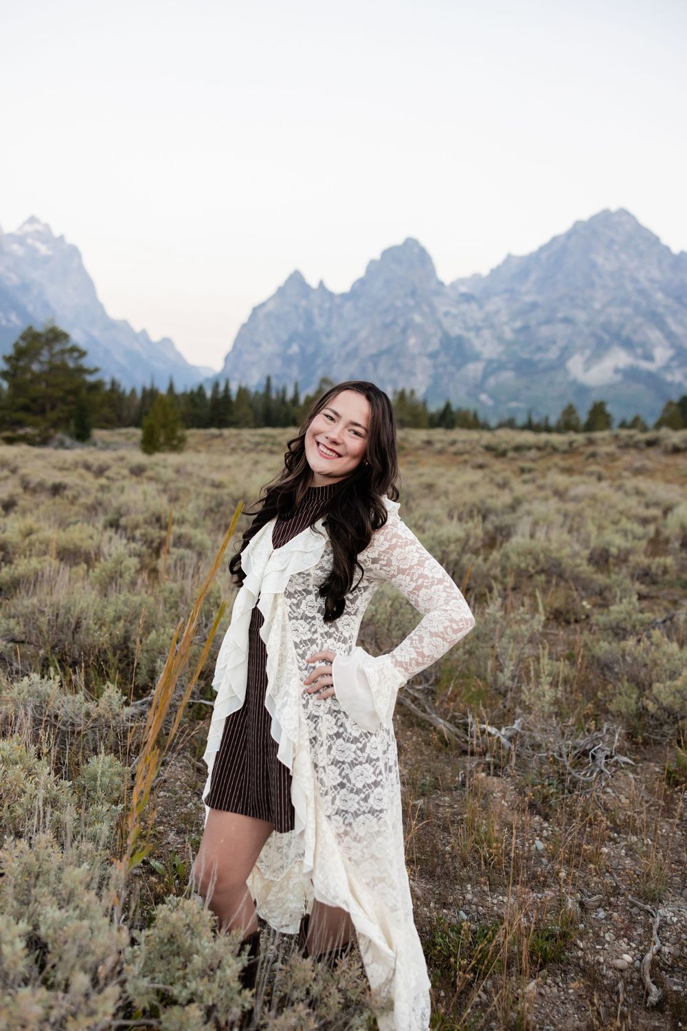 Young woman in white lace cardigan smiling in a field with Grand Teton mountains in the background.