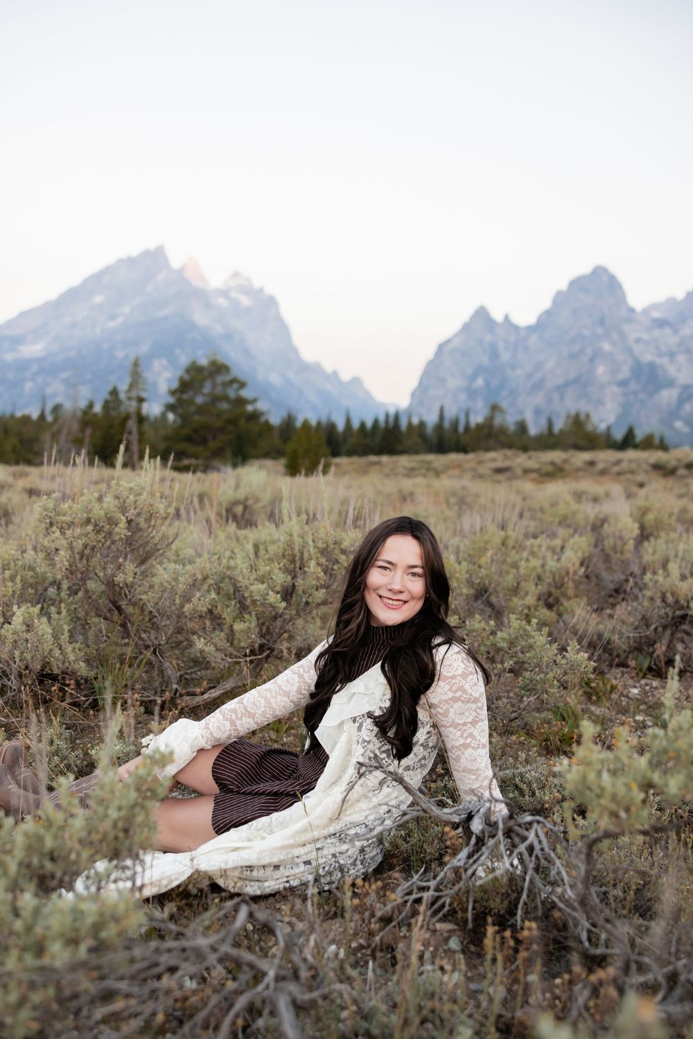 A smiling woman in a lace cardigan sits in a sagebrush meadow with the Grand Teton mountains in the background.