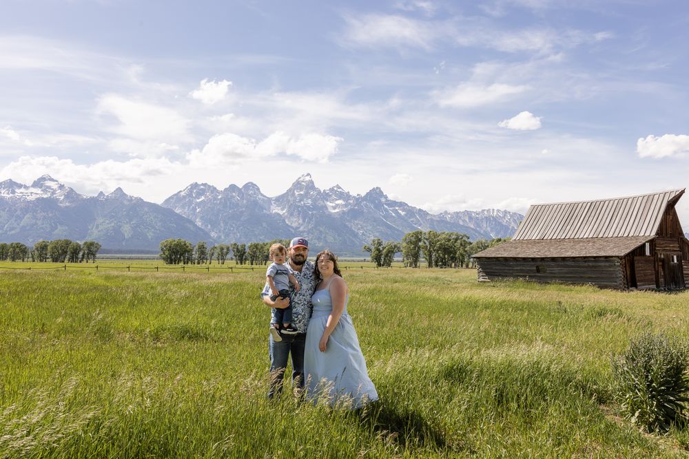 Couple with child poses in green meadow with Grand Teton mountains and rustic barn backdrop on wedding day.