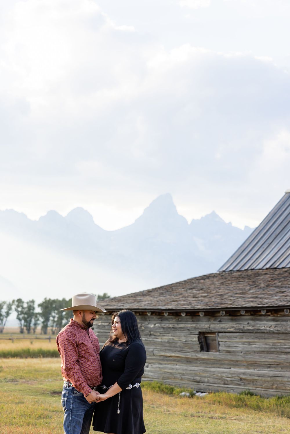 Couple holding hands before rustic barn with Grand Teton mountains backdrop, man in cowboy hat and woman in black dress.