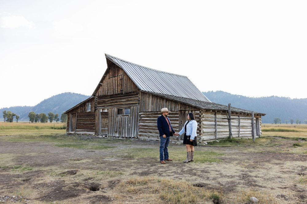 Two people stand before a rustic wooden barn in Grand Teton National Park with mountains in the background.