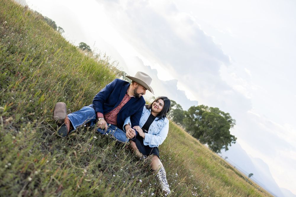Couple sitting together in a green meadow, man in cowboy hat and woman in denim jacket, sharing a romantic moment outdoors.