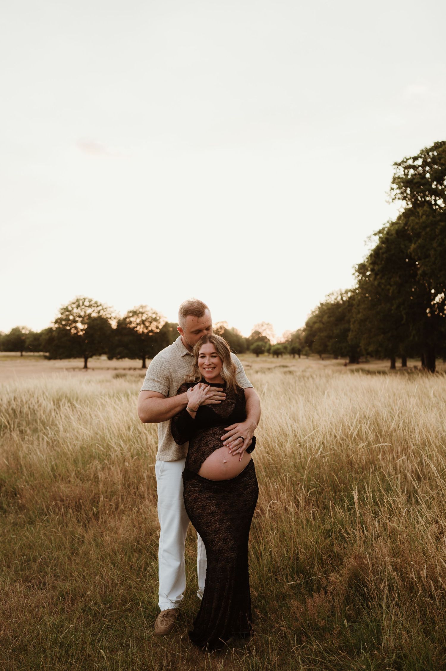 Couple embracing in golden field at sunset, pregnant woman in lace gown, man in white pants, trees in background.