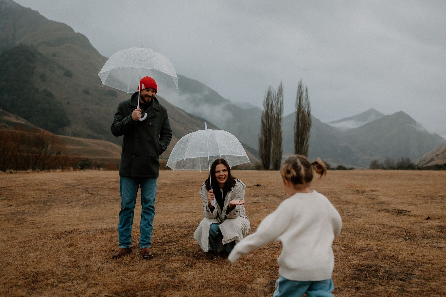Family with umbrellas enjoying a rainy day in a scenic mountain valley, parents smiling as child runs toward them.