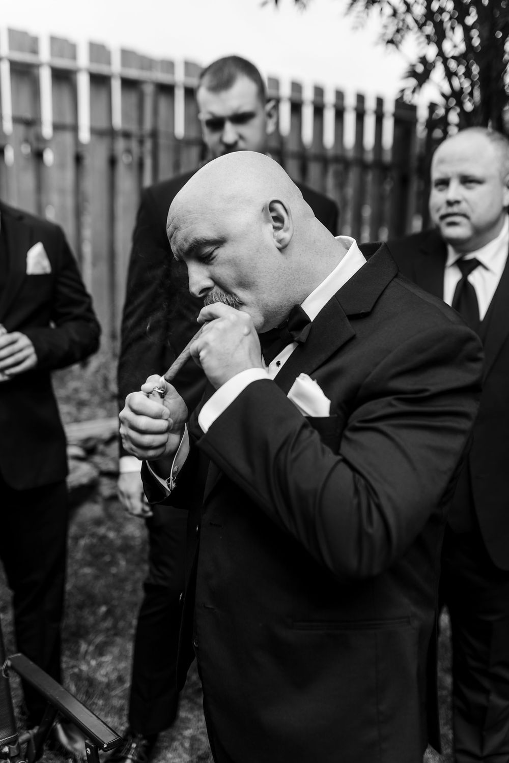 Bald groom in tuxedo lighting a cigar outdoors with groomsmen in black suits standing behind him.