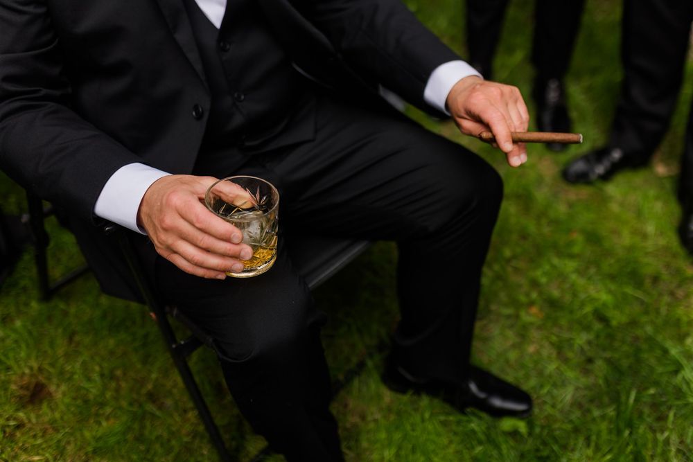 Man in black suit holding whiskey glass and cigar while seated outdoors on grass.