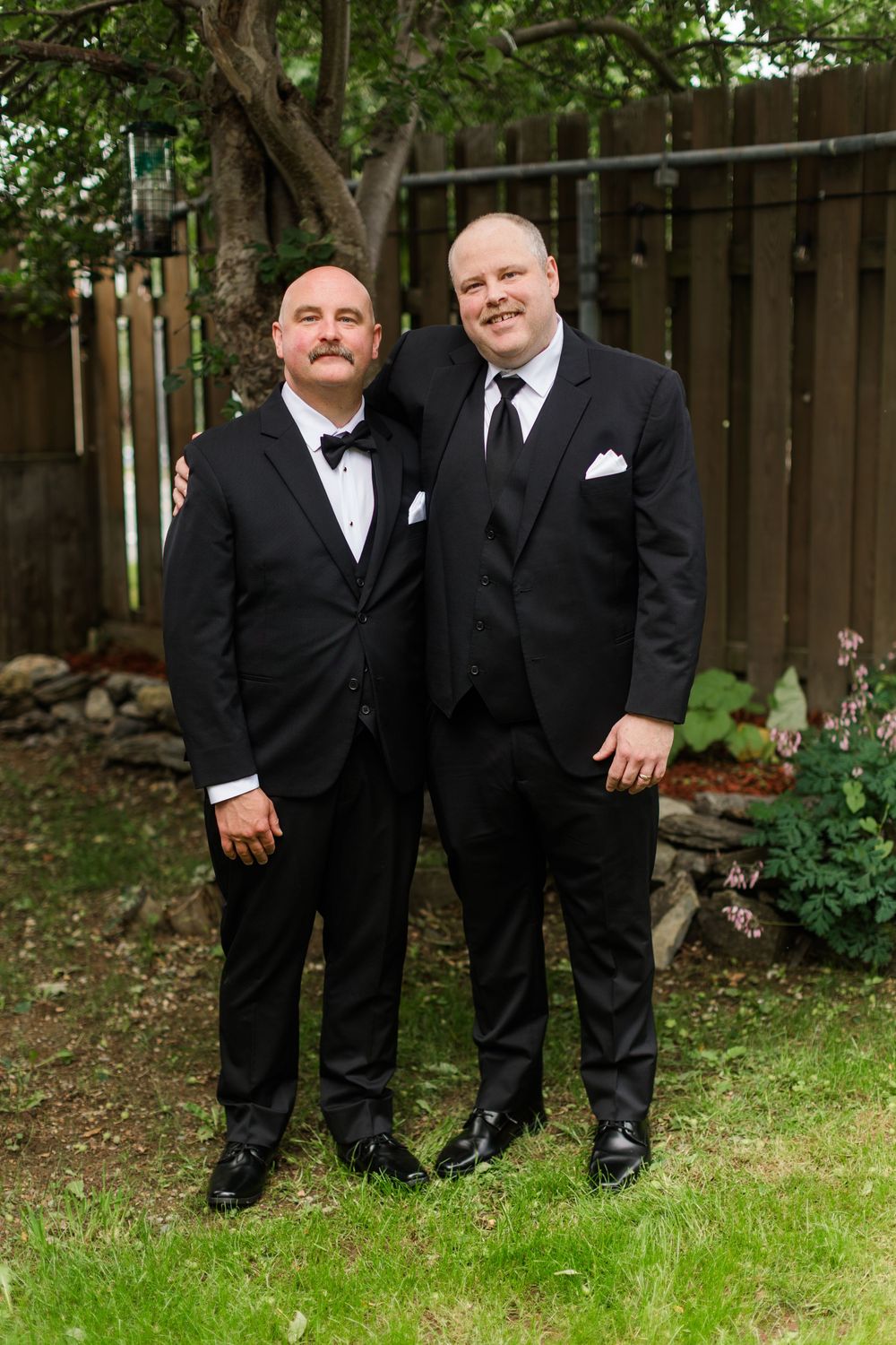 Two men in black tuxedos posing together outdoors at a wedding ceremony near a wooden fence.