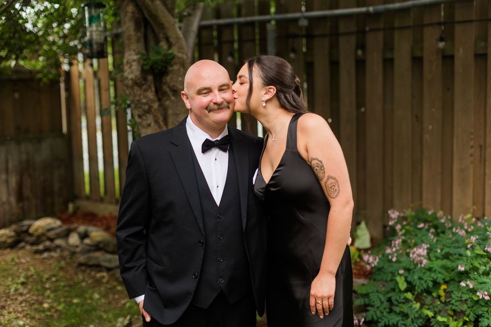 Couple in formal black attire sharing a tender kiss in a garden, man in tuxedo and woman with tattoo.