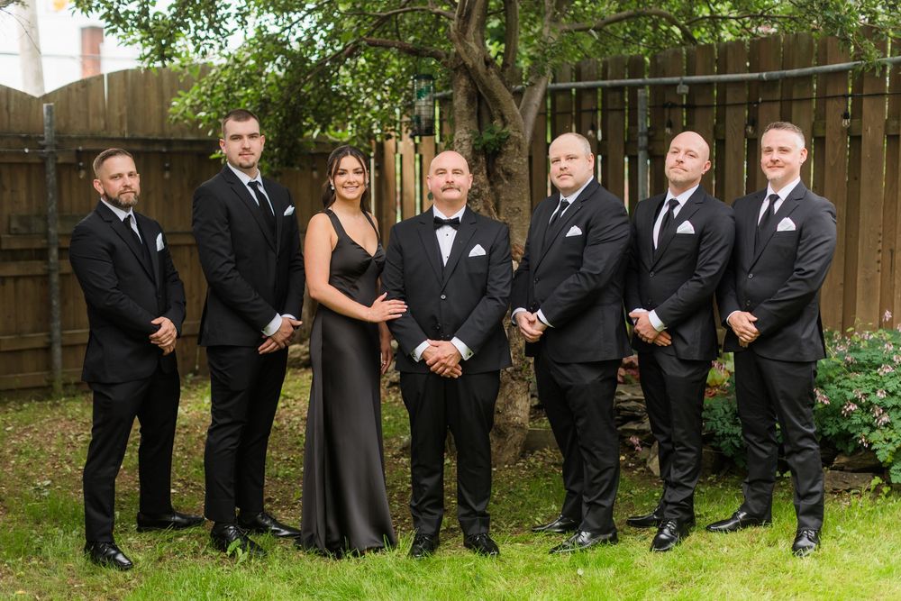 Wedding party in black formal attire posing outdoors, groom in tuxedo surrounded by groomsmen and bridesmaid.
