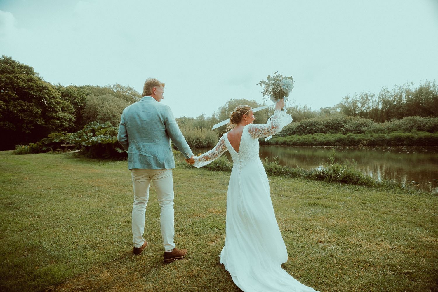 A bride and groom hold hands while walking toward a serene pond surrounded by lush greenery on their wedding day.