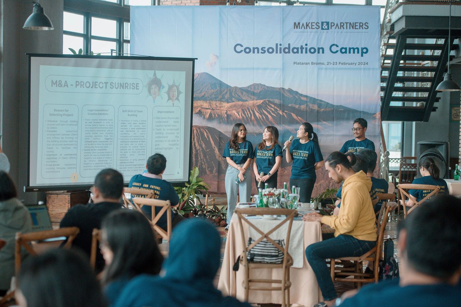 Group of professionals presenting at a Makes & Partners Consolidation Camp event with a mountain backdrop banner.