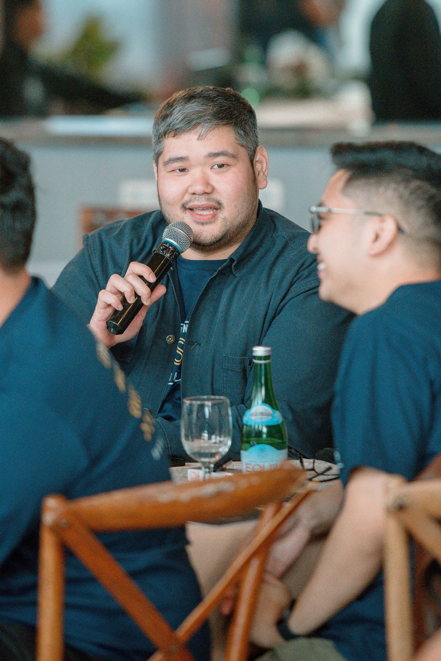Man speaking into microphone at restaurant table with green bottle and wine glass nearby.