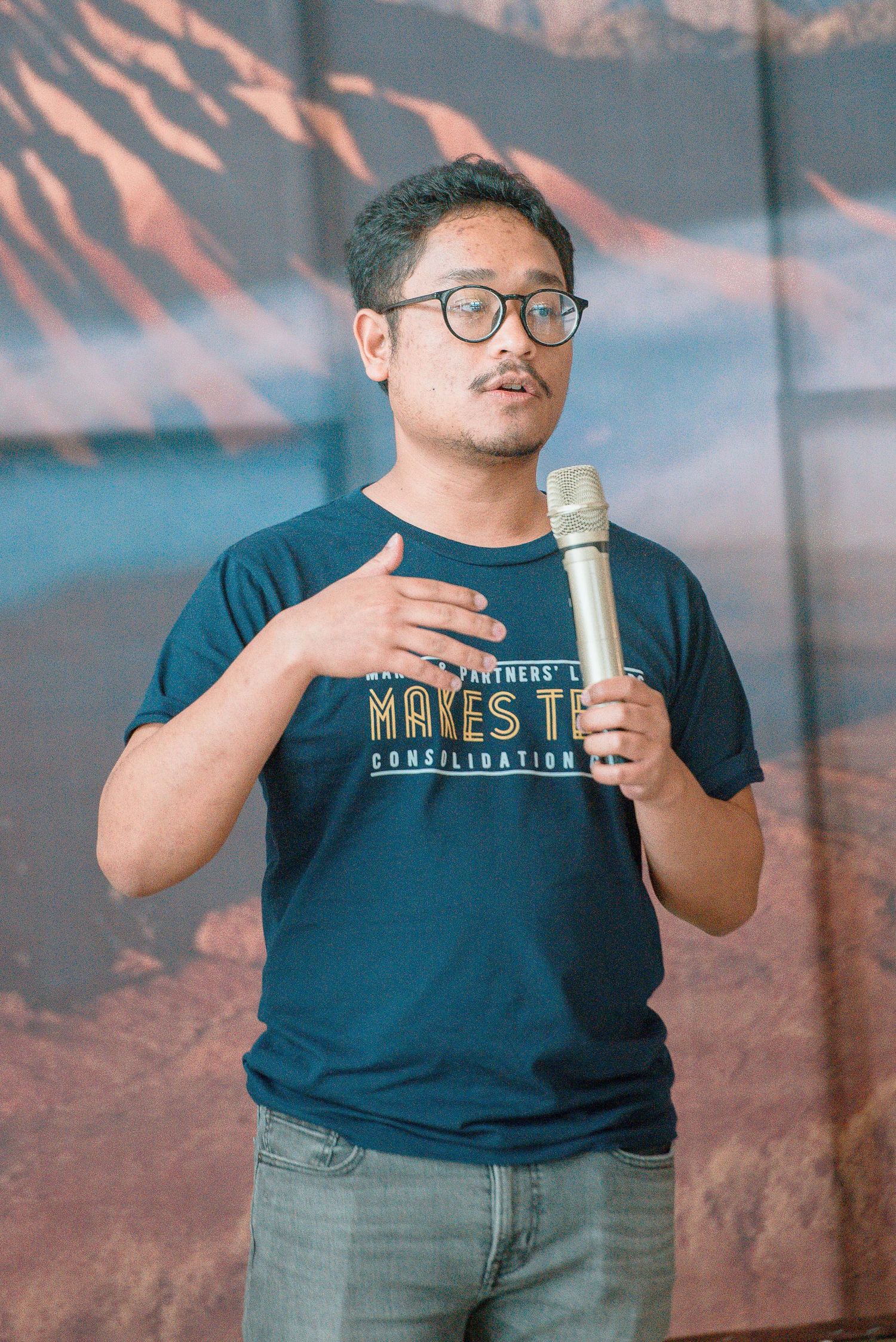Man in glasses and navy t-shirt speaking into microphone at outdoor event.