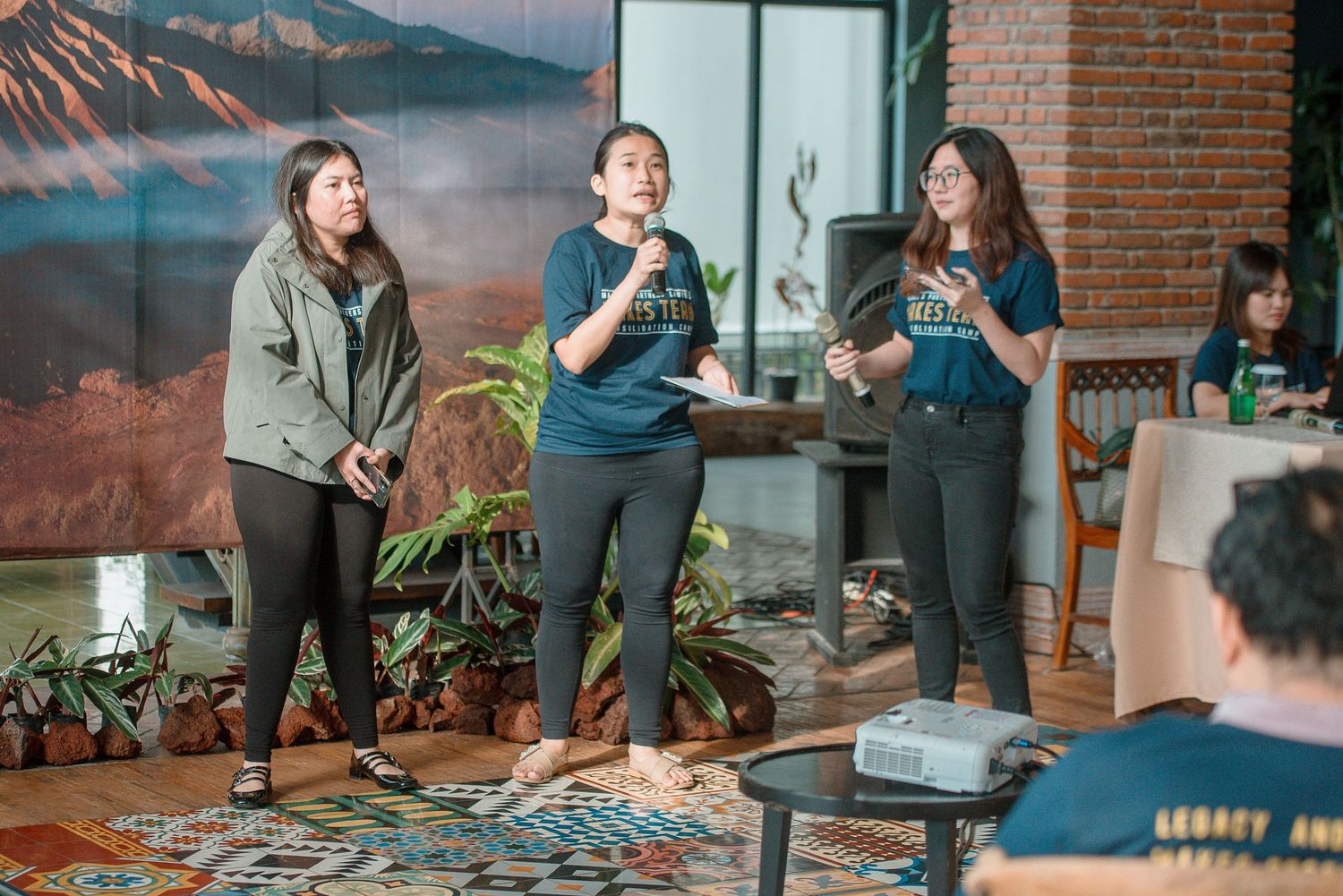 Three women presenting indoors with tropical decor, plants, and a mountain mural backdrop at a casual event.
