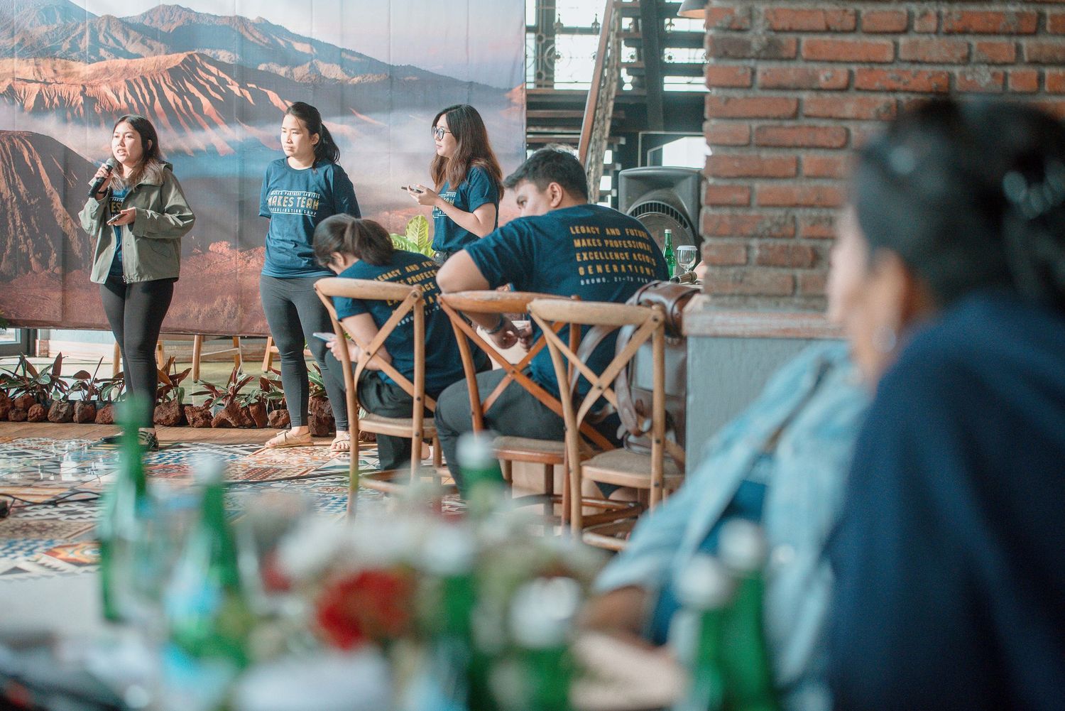 People dining at a rustic restaurant with brick walls, bamboo furniture, and a mountain mural backdrop.