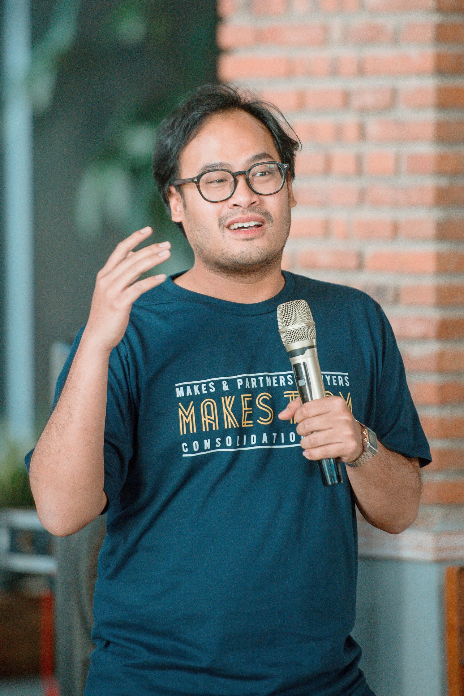 Man in dark teal graphic t-shirt speaking into microphone, gesturing with hand, wearing glasses, brick wall background.