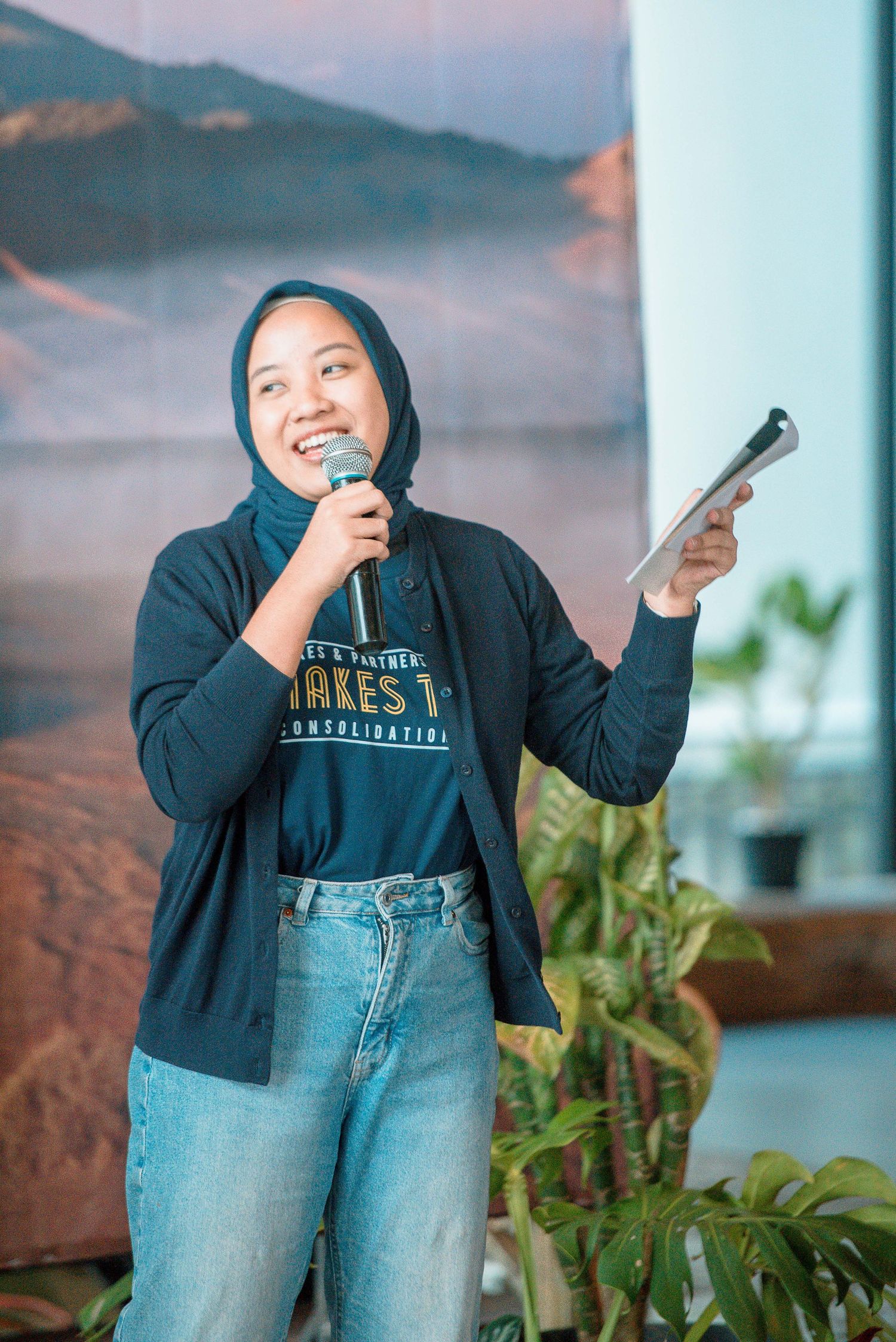 A young woman in hijab and casual attire speaks into a microphone while holding a tablet at an indoor event.