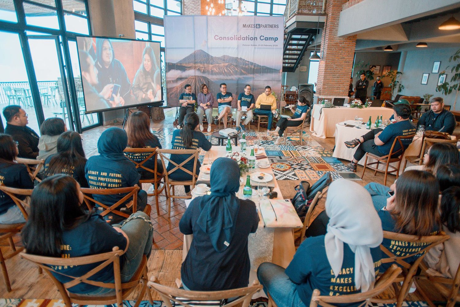 Group of diverse attendees engaged in a panel discussion at an indoor community camp event with a projected screen.