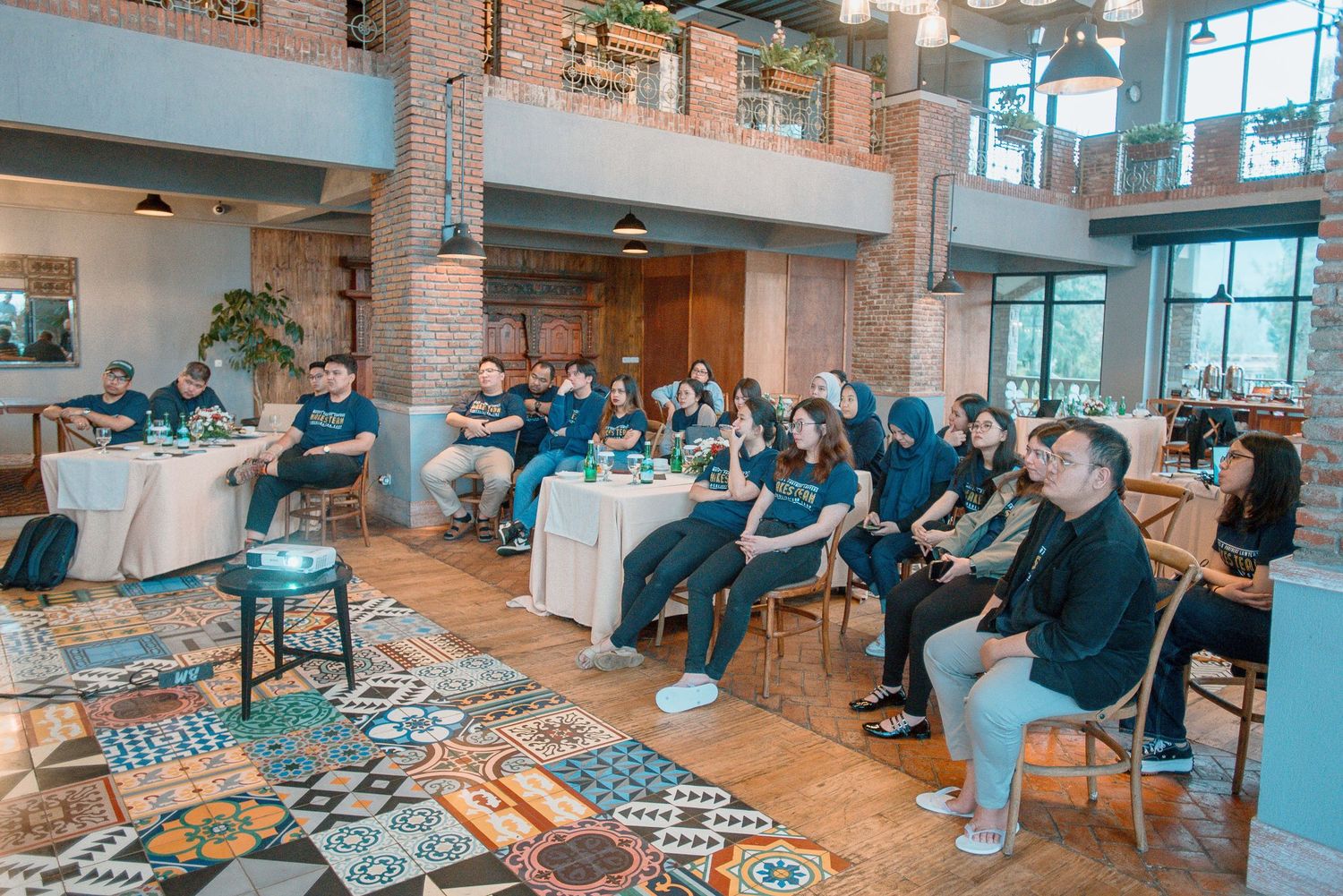 Group of people seated in a stylish cafe with colorful tile floors, brick walls, attending a casual indoor event.