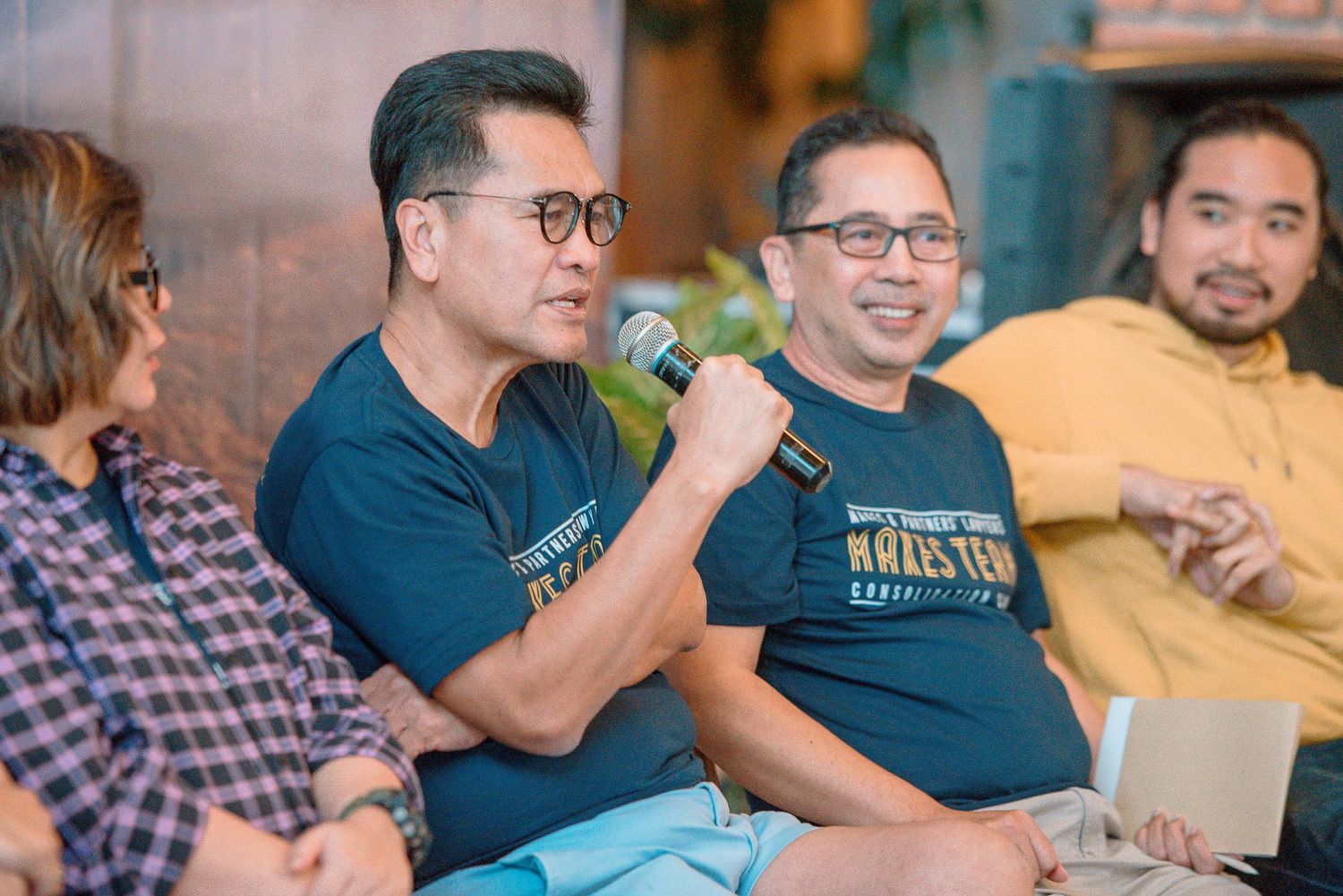 Group of men in casual blue shirts smiling and speaking into a microphone at an indoor panel discussion event.