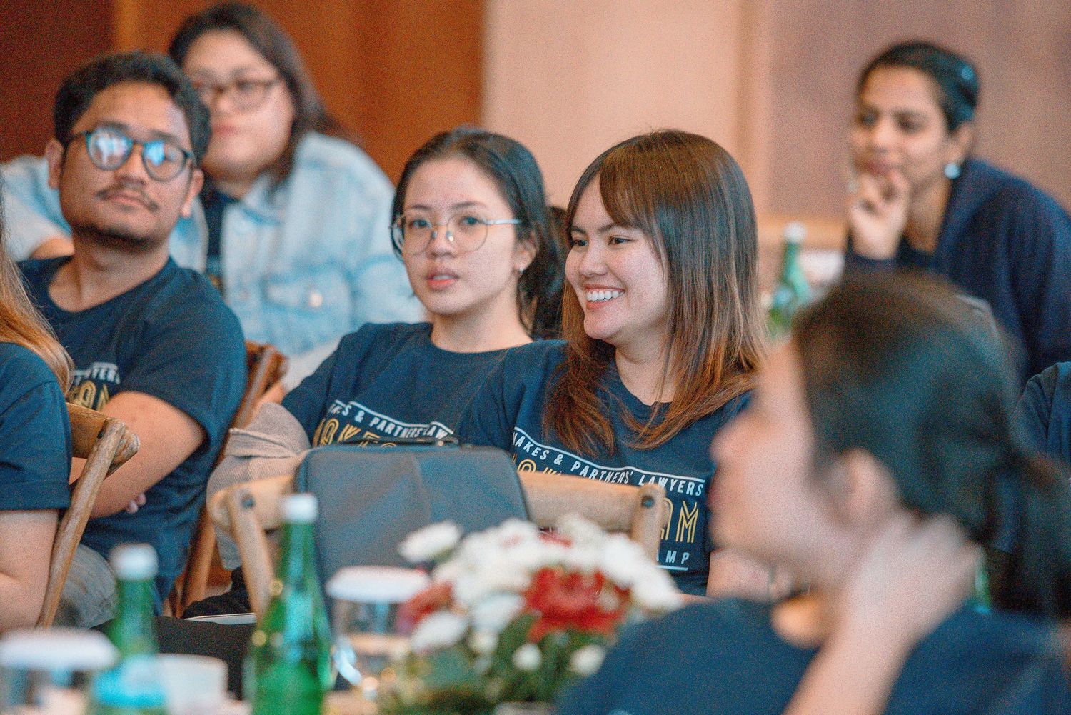 Group of young students in matching navy shirts smiling and engaged at a conference table with flowers and drinks.