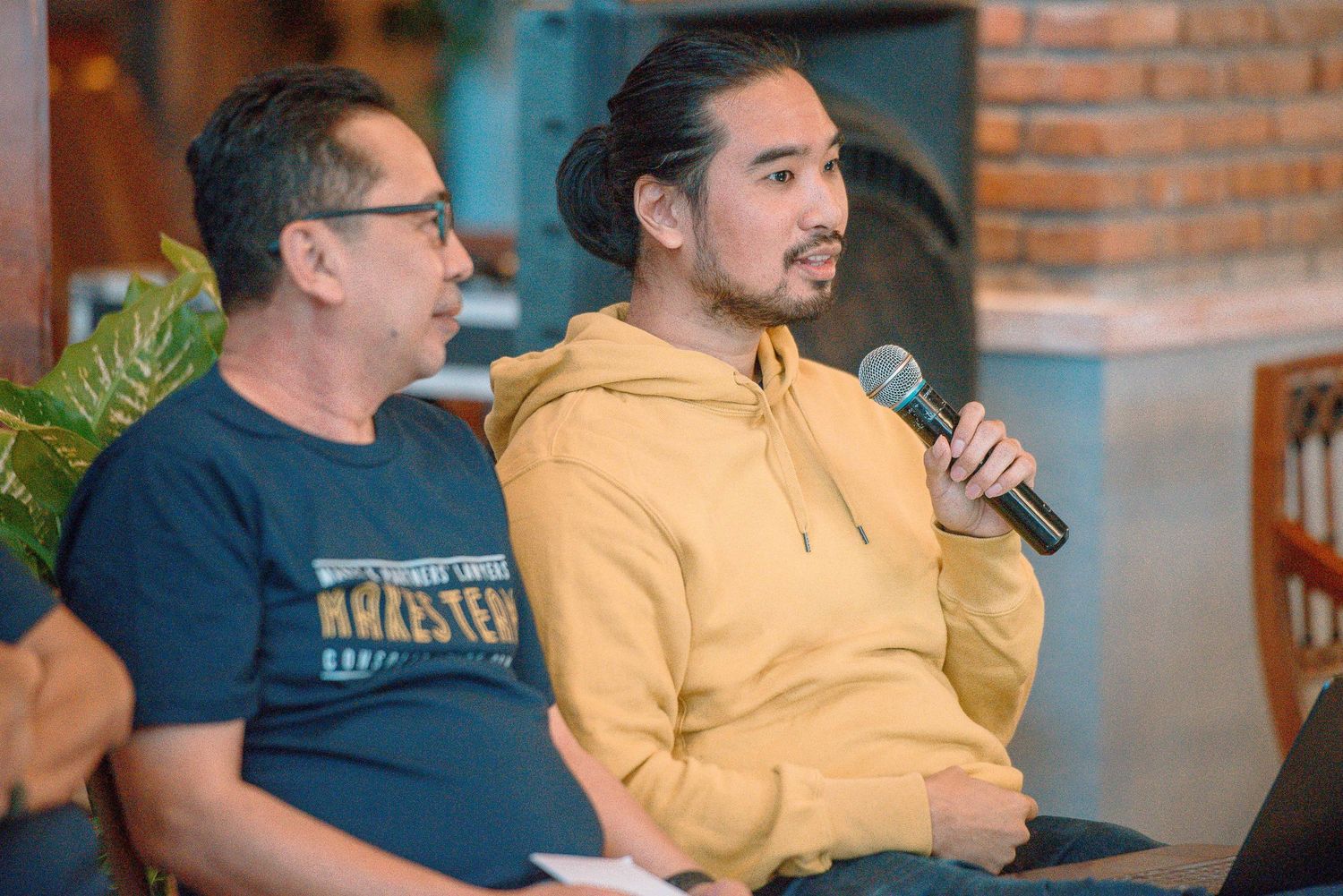 Two men sit together, one speaking into a microphone wearing a yellow hoodie, at an indoor casual panel discussion.