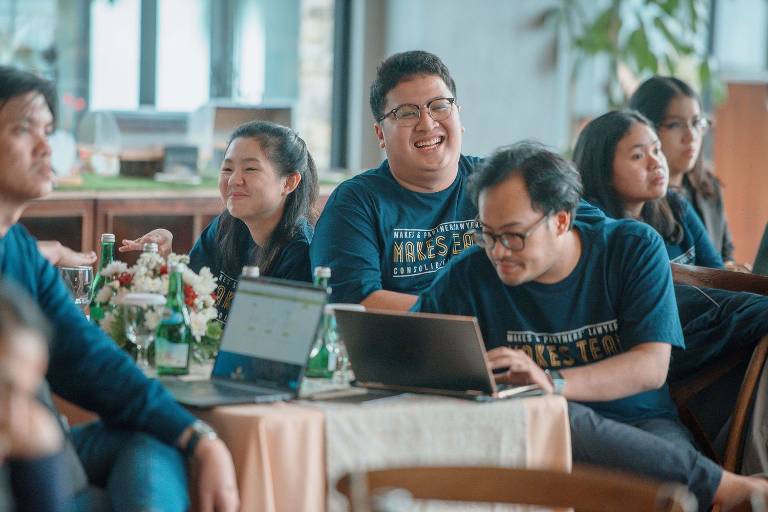 Group of colleagues in matching teal shirts smiling and working on laptops during a team meeting.