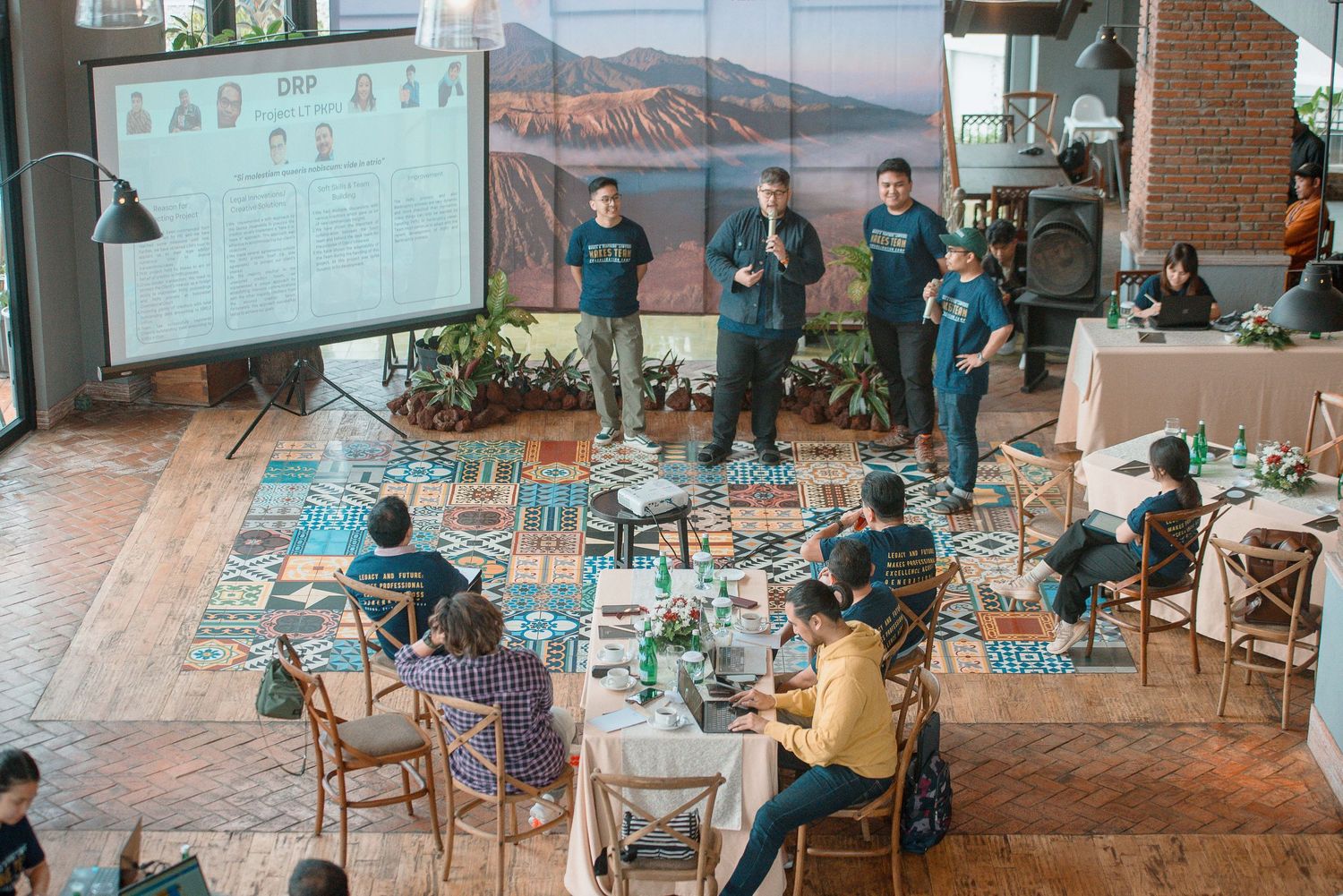 A group presentation at an indoor event with speakers on a patterned floor, audience seated at tables, and a large screen display.