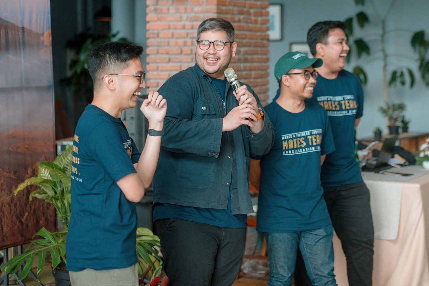 Four men in matching teal shirts laugh together at an indoor event, one holding a microphone.