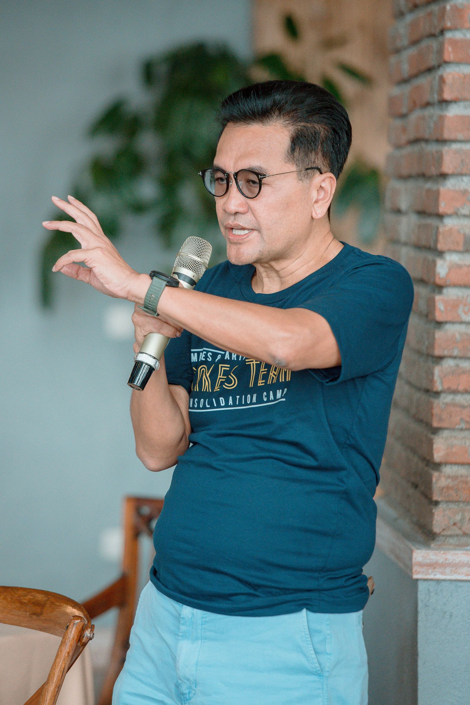 Man in teal graphic t-shirt holding microphone and gesturing while speaking at an indoor event.