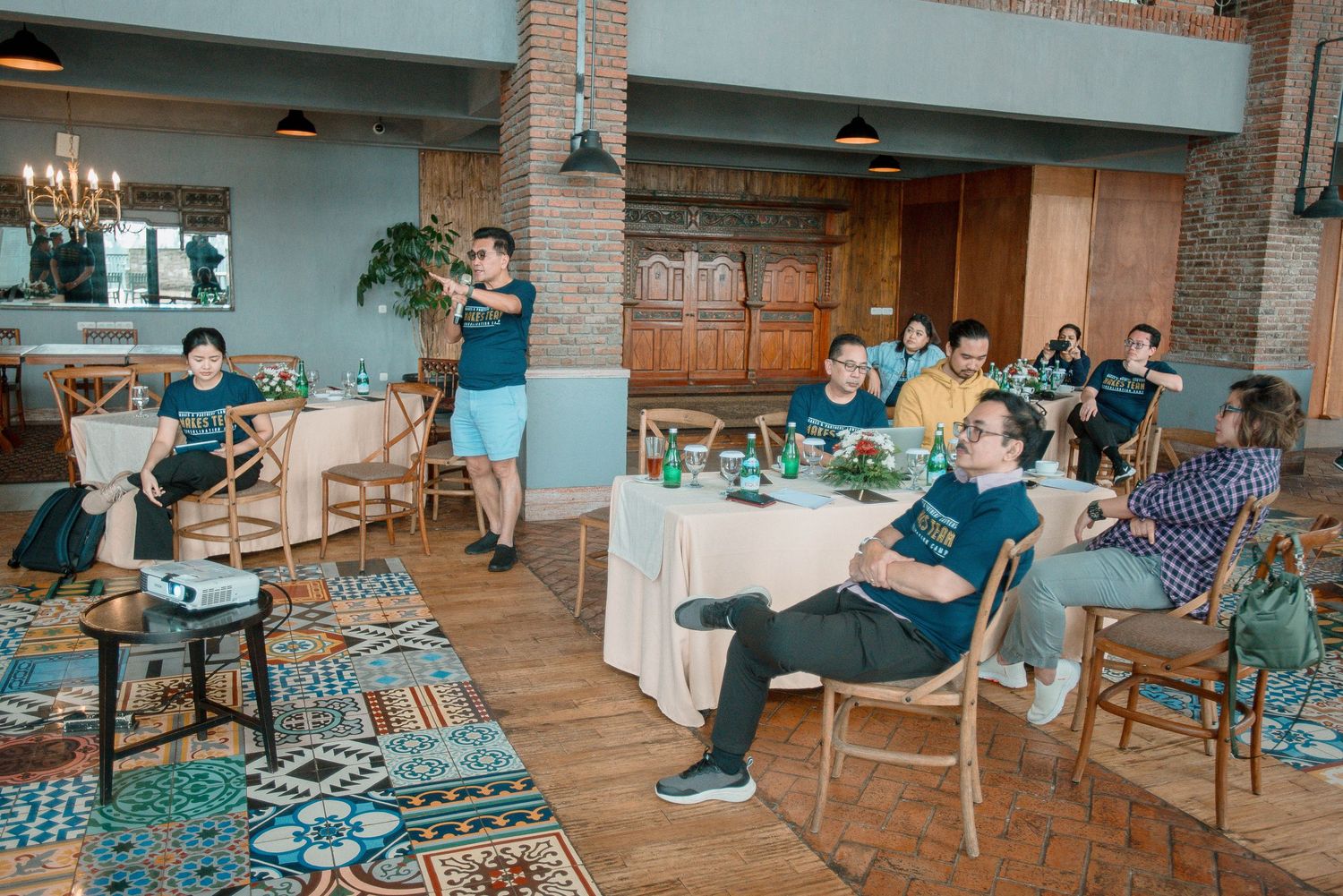 Group of people seated at tables in a rustic restaurant watching a presentation on a projector screen.