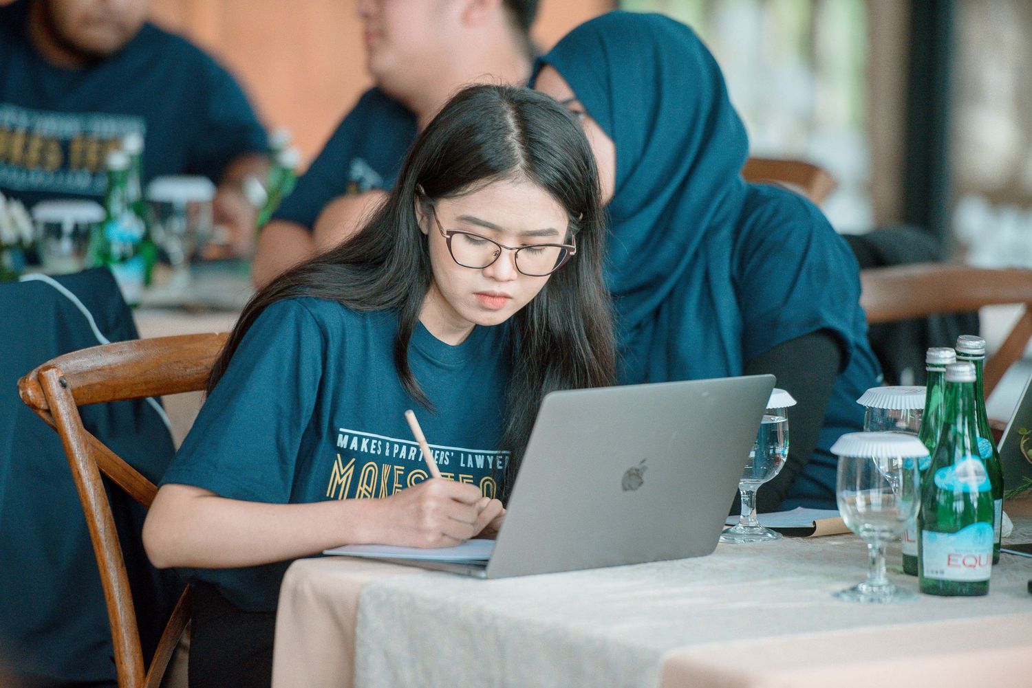 Young woman with glasses works on laptop at table during group event, wearing teal shirt.
