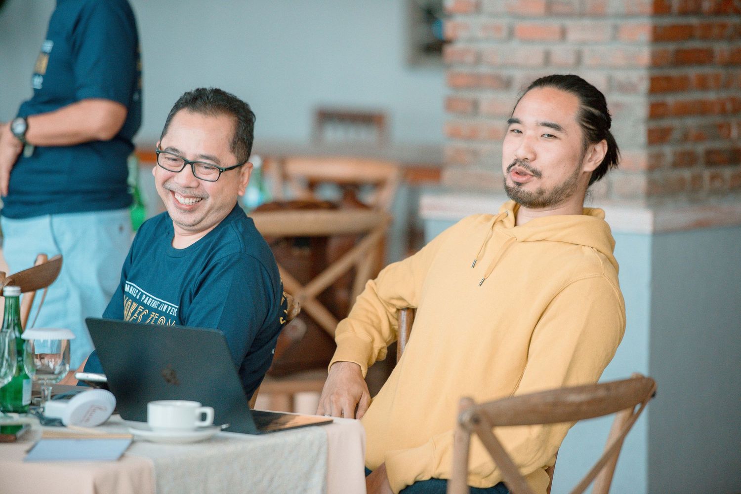 Two smiling Asian men sitting at a cafe table with a laptop and coffee cup in a modern restaurant setting.