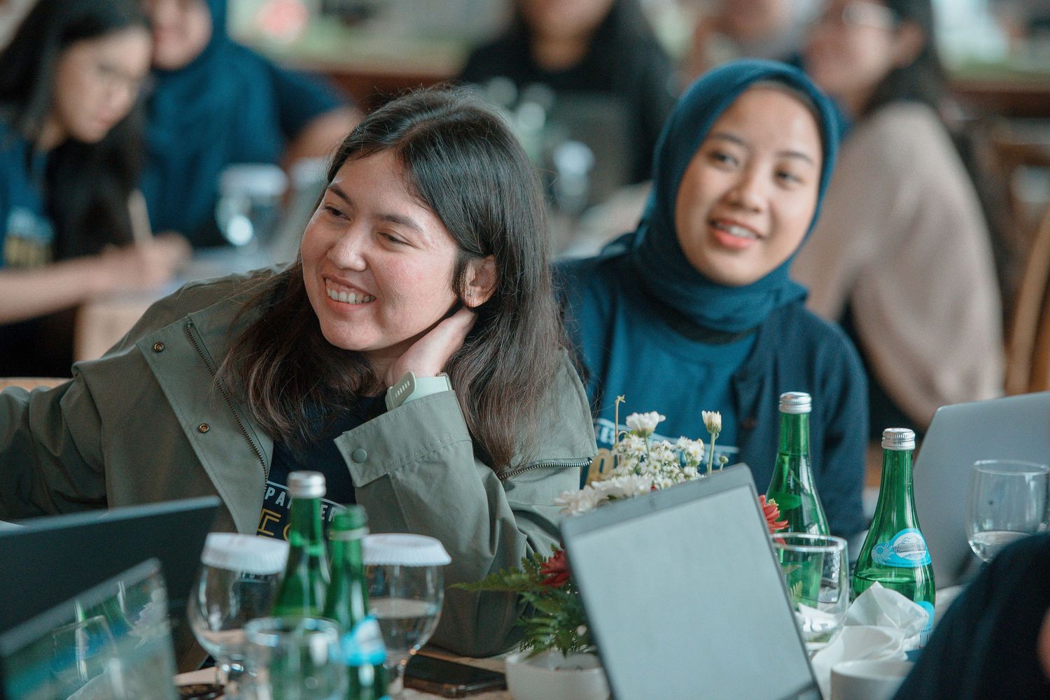 Two women smiling at a conference table with laptops and water bottles, one wearing a hijab.