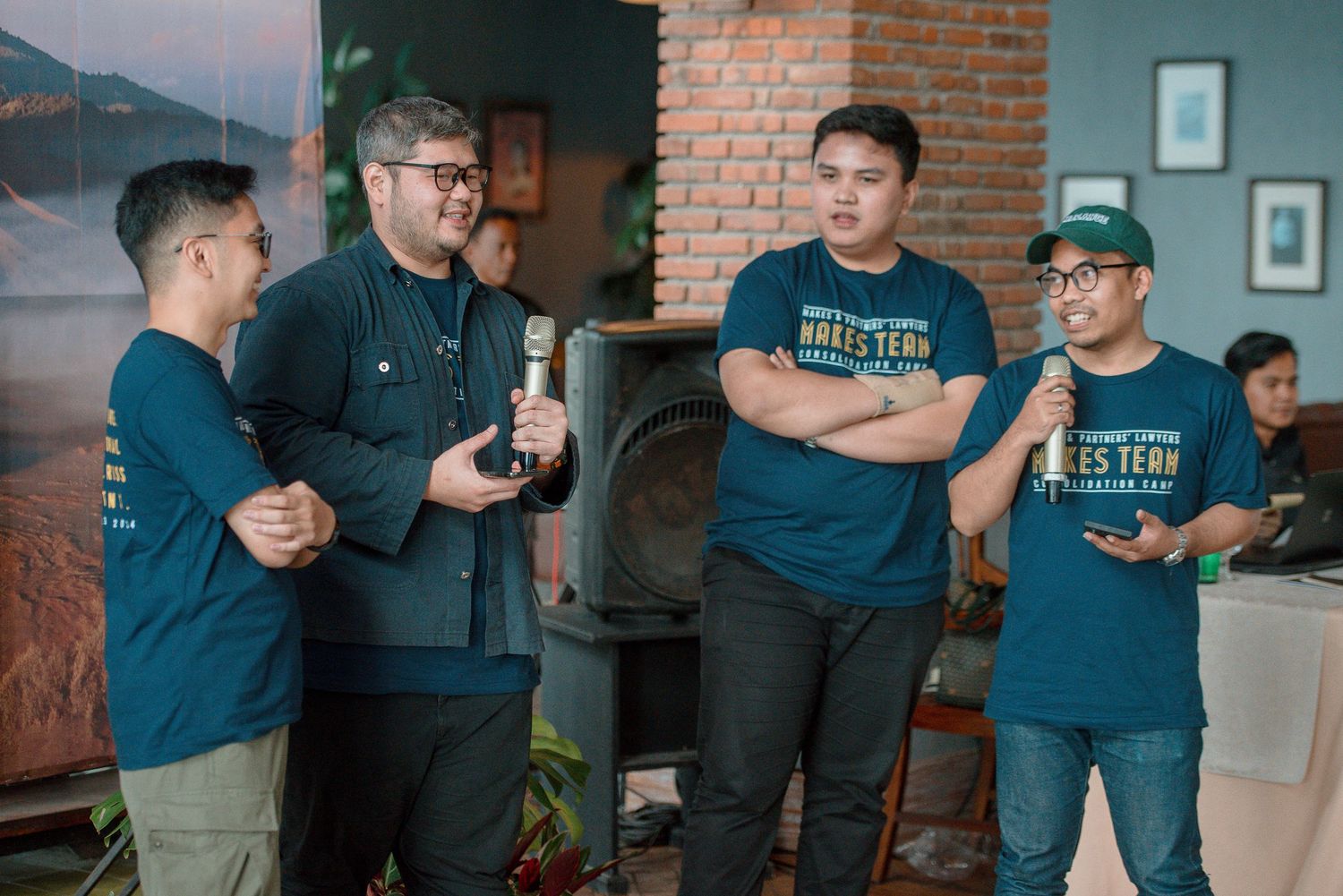 Four men in matching teal shirts stand together at an indoor event, one holding a microphone during a presentation.