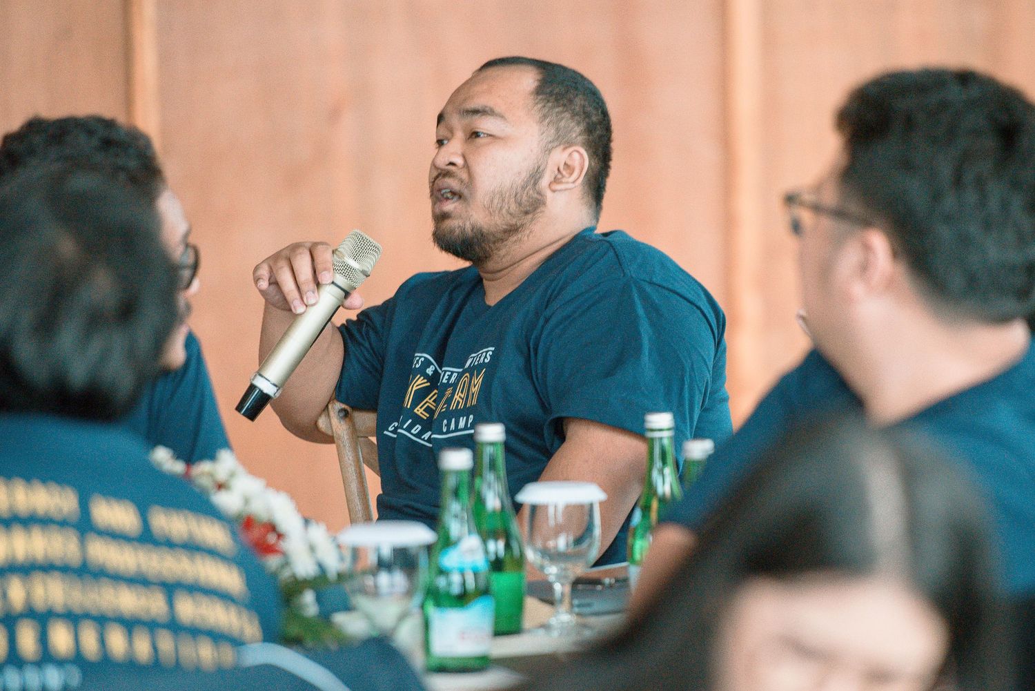 Man speaking into microphone at conference table during group discussion, wearing teal shirt.
