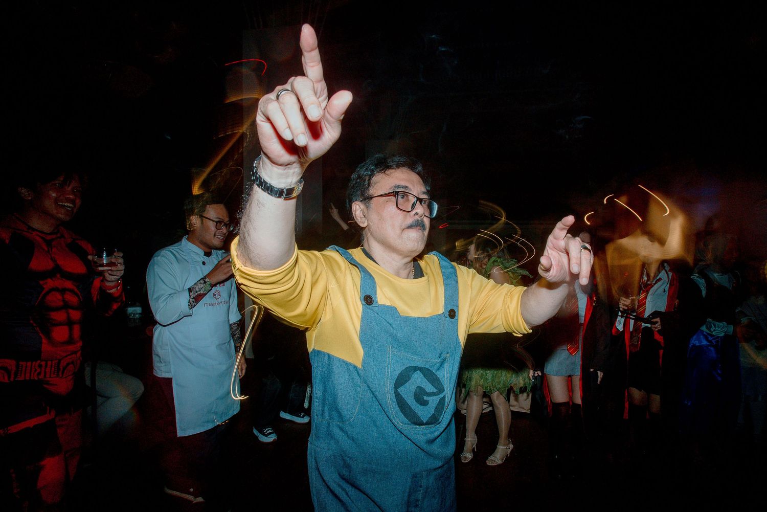 Person in Minion costume dancing at a Halloween party with colorful crowd in background.