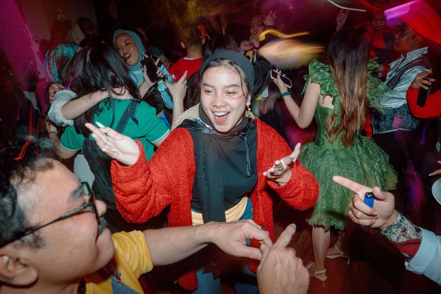 People in colorful costumes dancing energetically at a festive Halloween party with vibrant pink lighting.