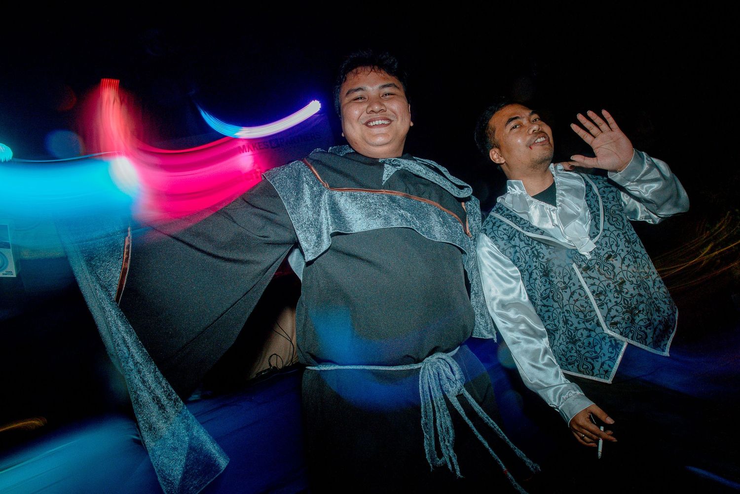 Two people in ornate silver costumes smile and wave against a dark background with colorful light streaks.