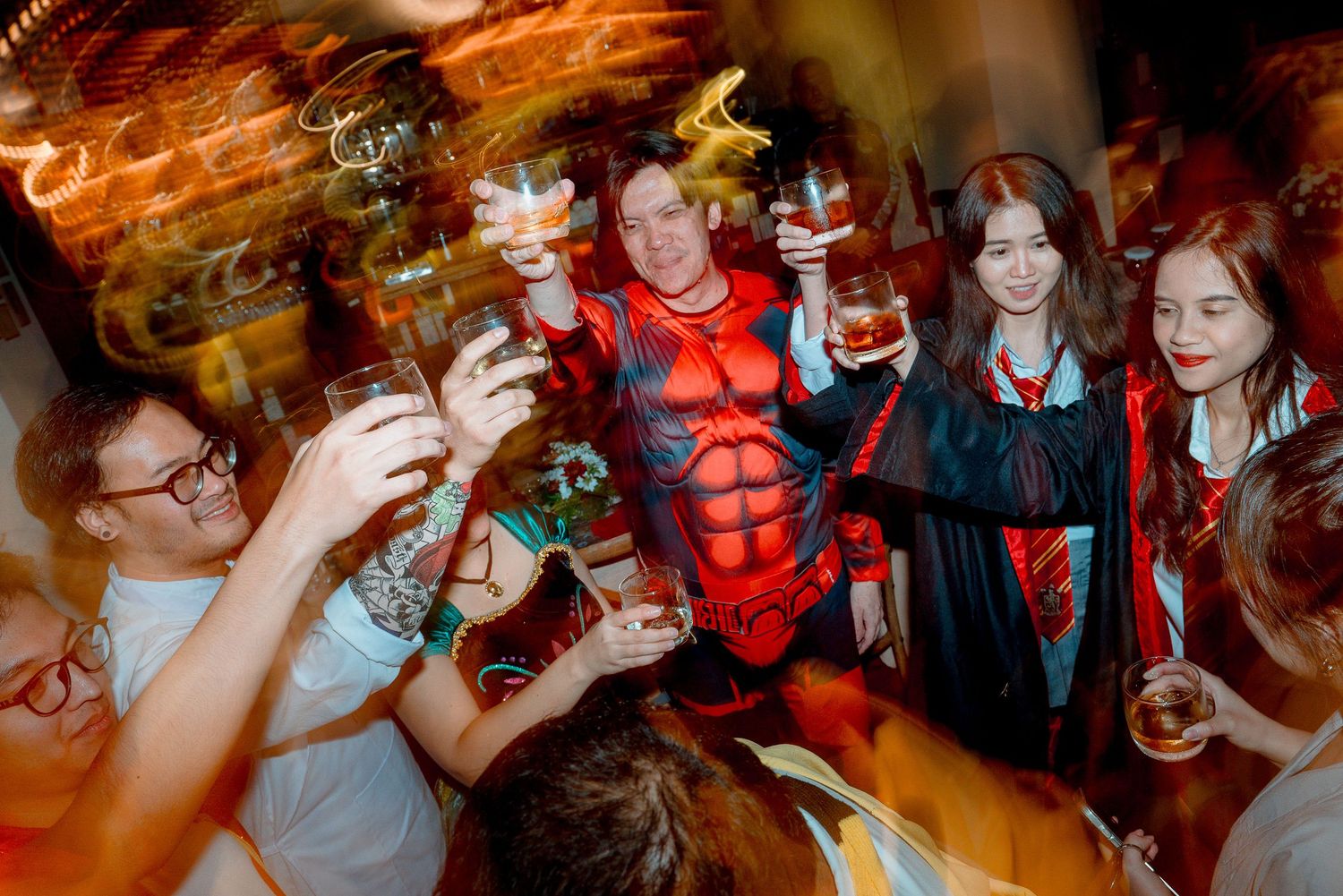 Group of people in Halloween costumes raising glasses in a toast at a festive party celebration.