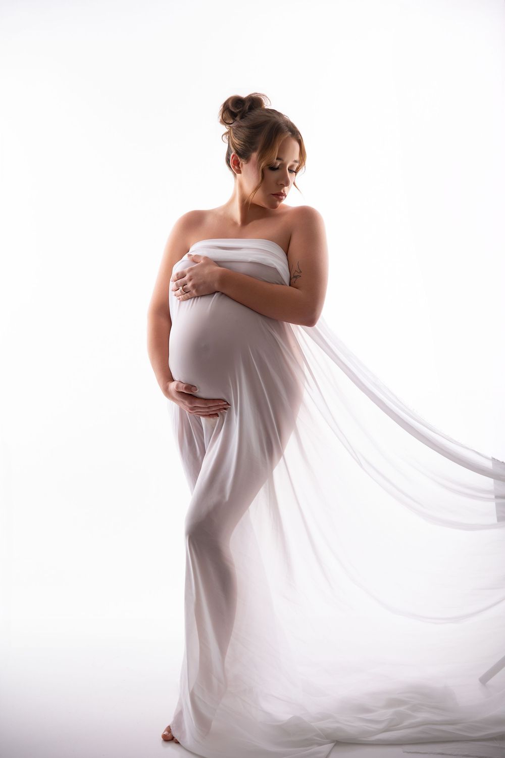 Pregnant woman draped in flowing white sheer fabric, cradling her baby bump in a serene studio maternity portrait.