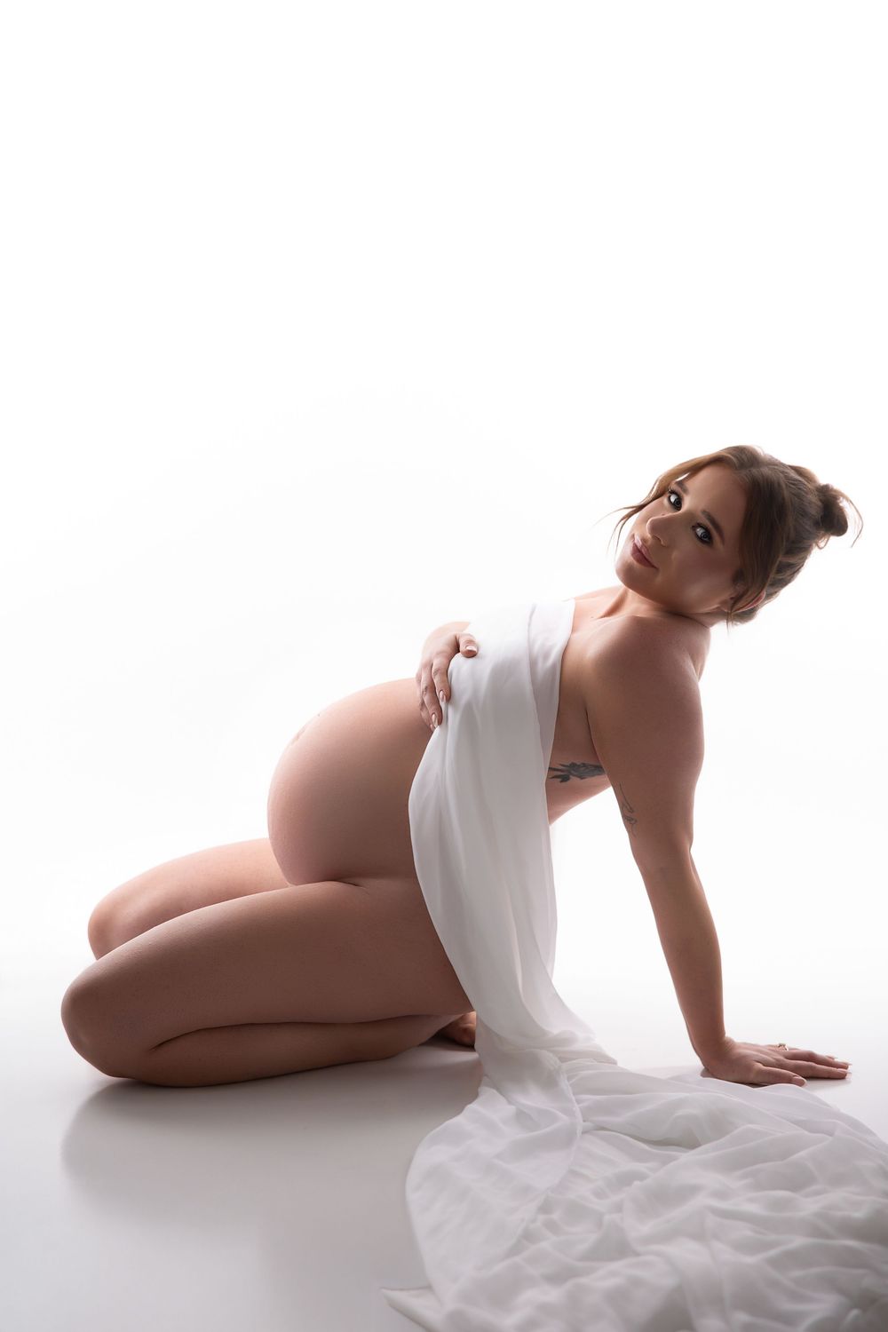 Pregnant woman kneeling on white floor draped in sheer white fabric, cradling her baby bump in a studio maternity photo.