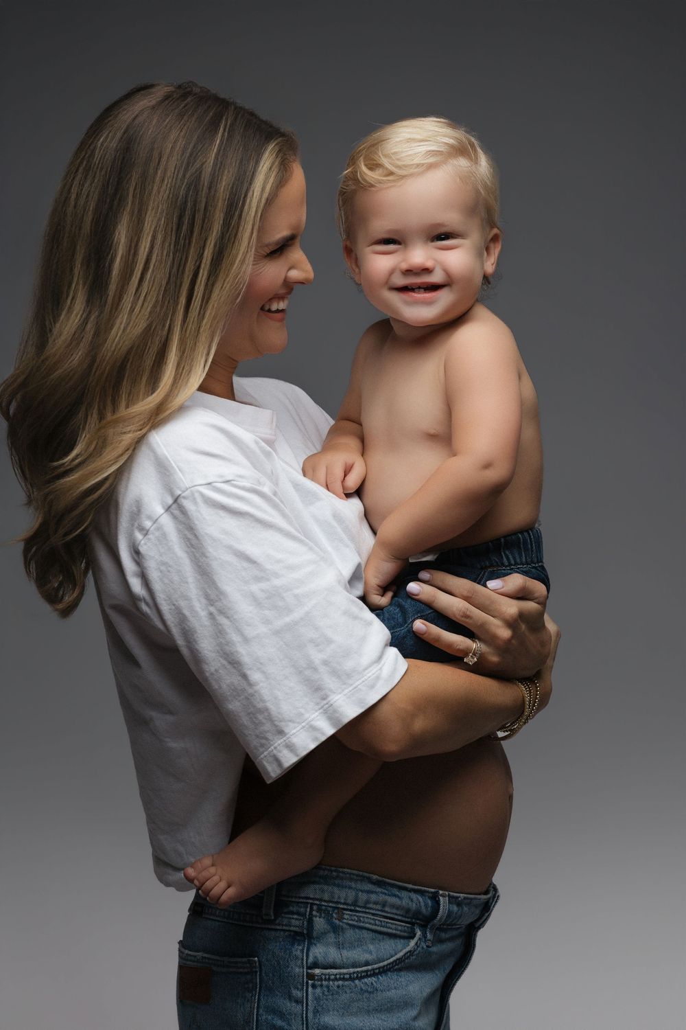 Smiling pregnant woman in white crop top holding happy shirtless toddler boy against gray background.