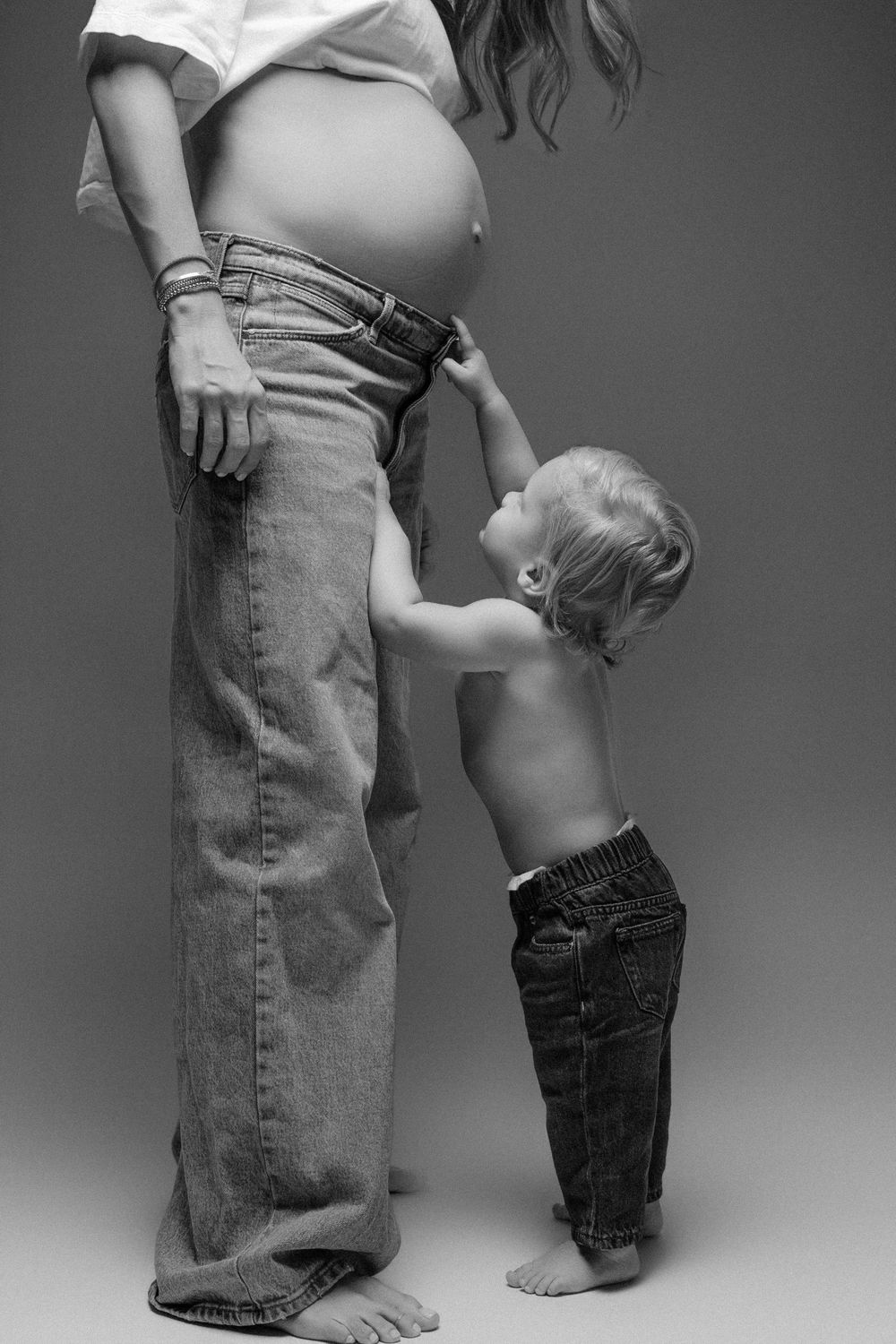 Black and white photo of pregnant woman with toddler boy touching her baby bump, both wearing jeans.
