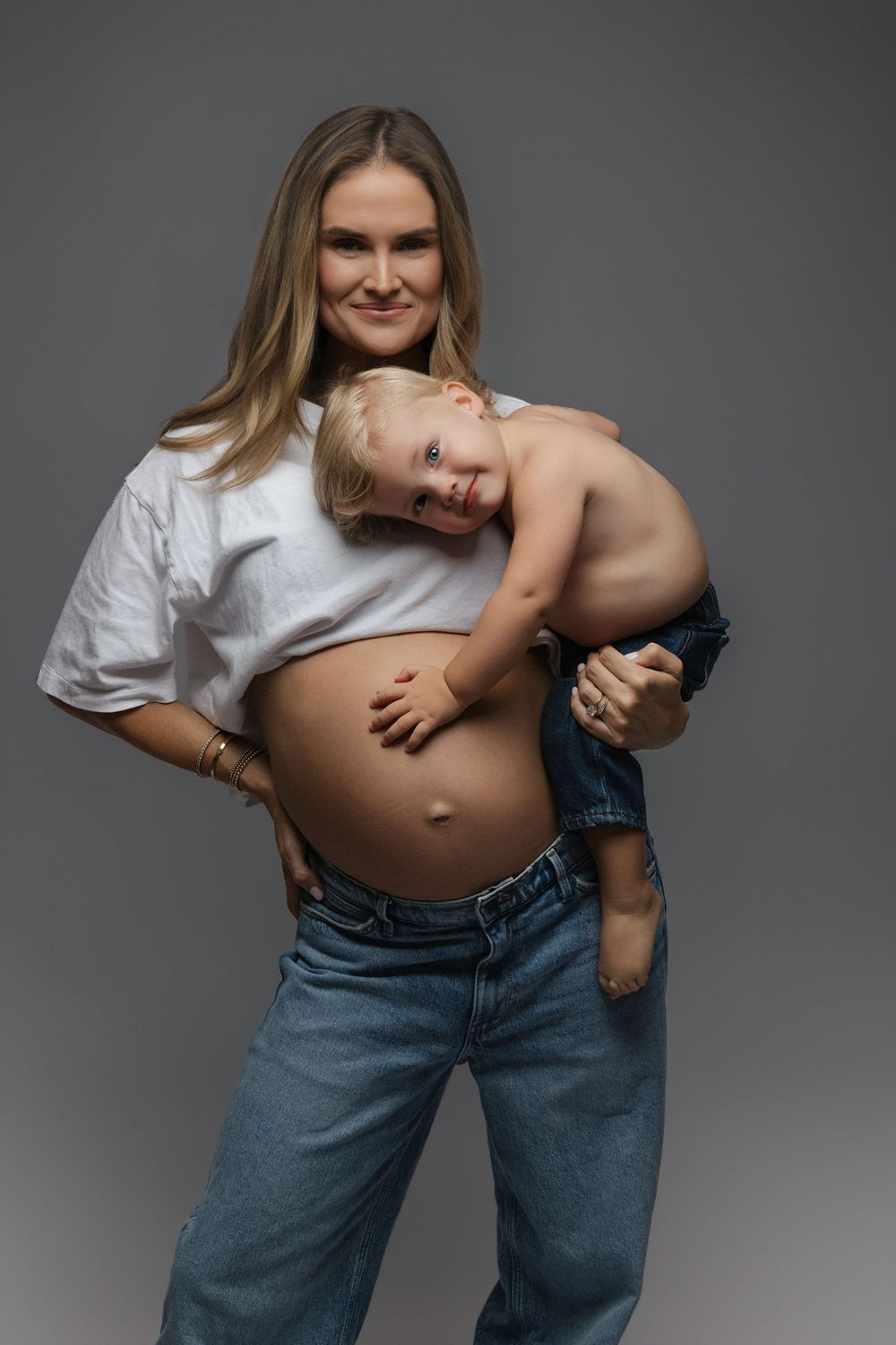 Pregnant woman in white crop top and jeans holds toddler child against her bare baby bump in studio.